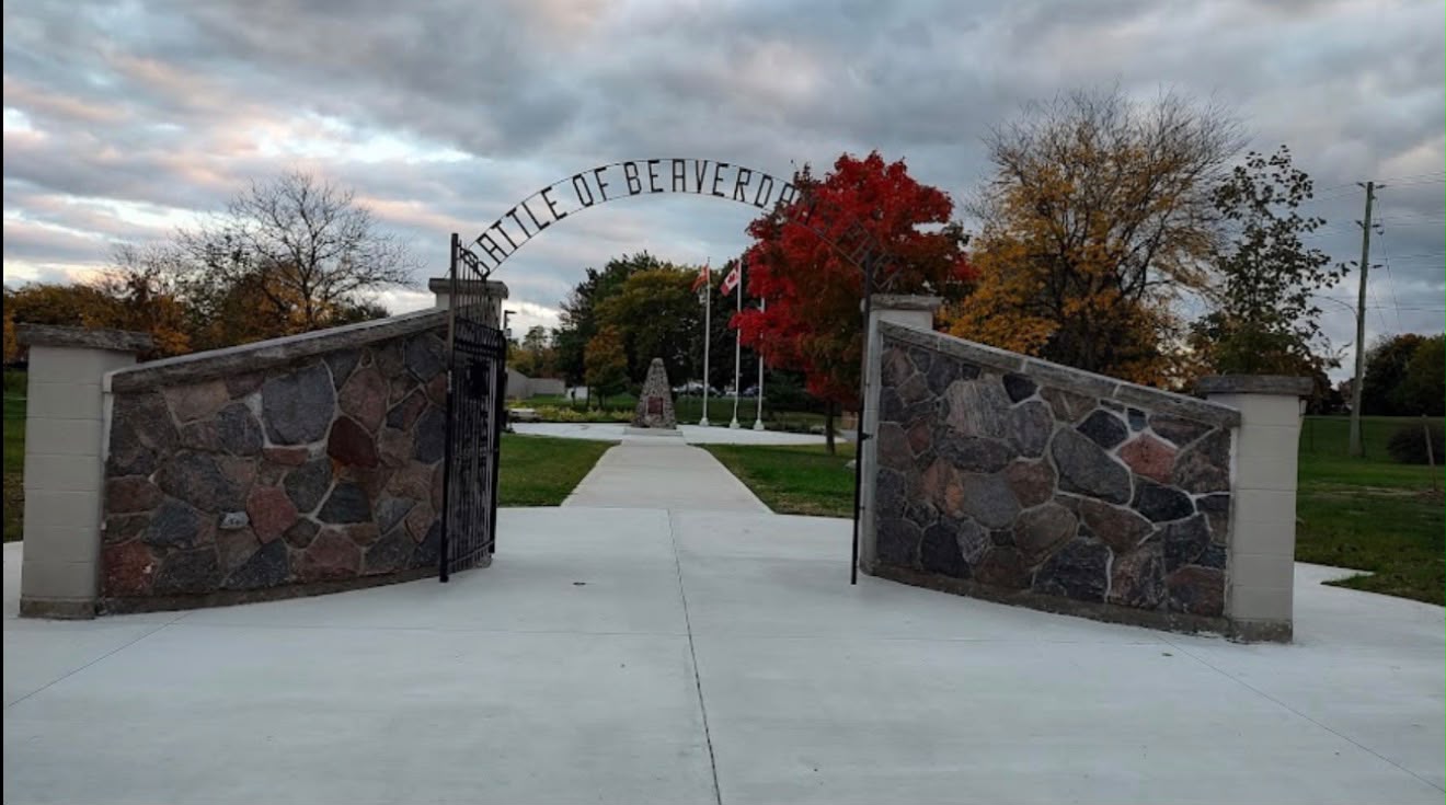 Stone gateway with open iron gates, “BATTLE OF BEAVER…” arch, autumn trees, flags, and a park walkway.