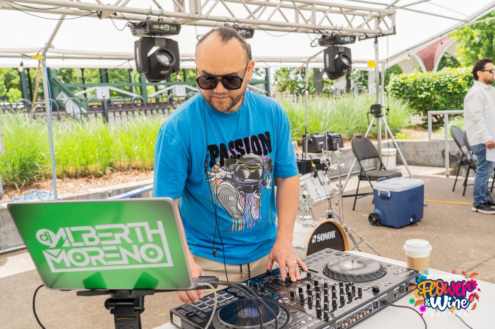 DJ in sunglasses performs under an outdoor canopy beside a laptop, stage lights, and drum equipment.