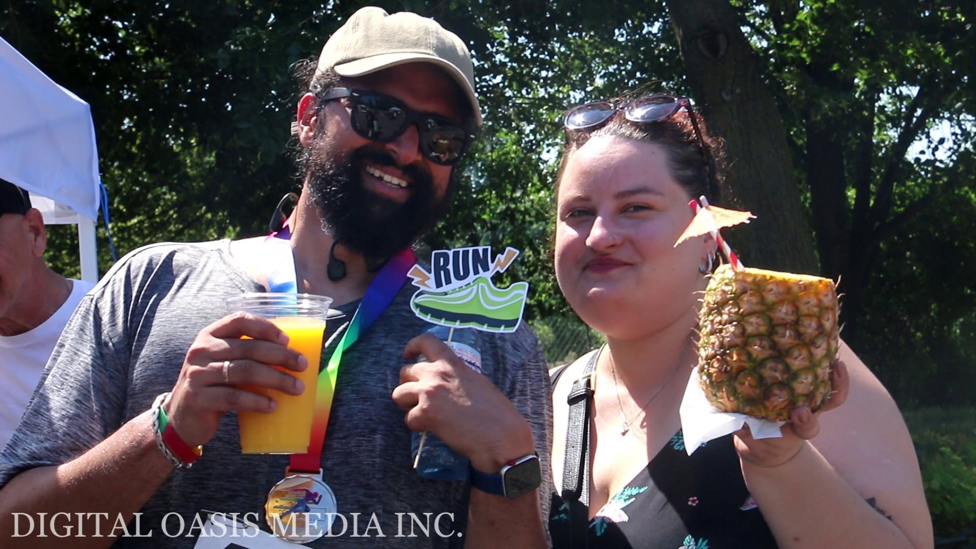 Two smiling event attendees hold orange juice and a pineapple drink beneath trees.