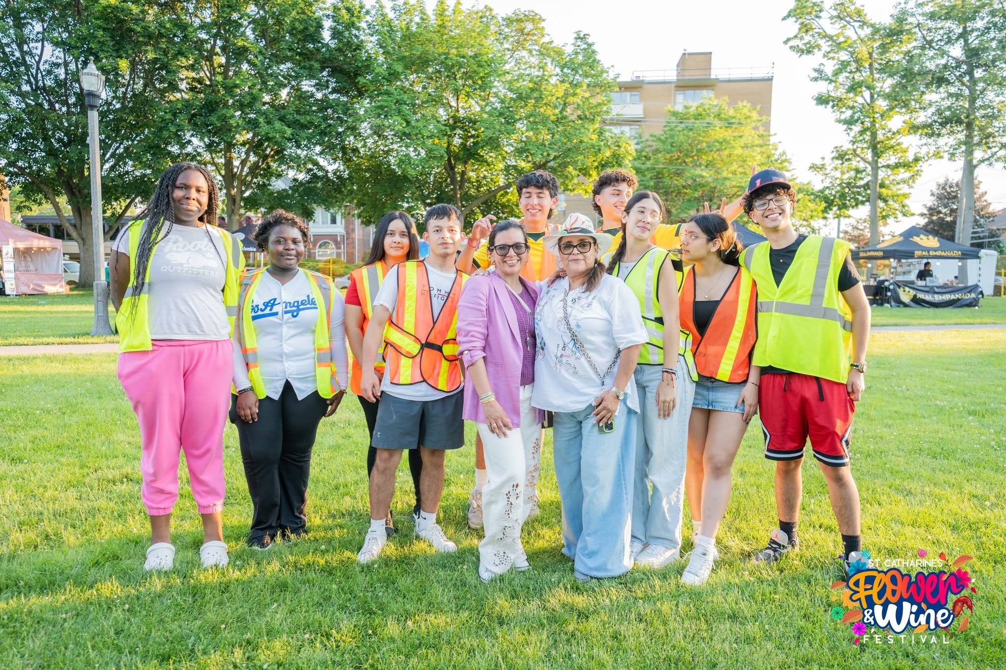 Diverse group in safety vests poses on a grassy festival lawn beneath trees.